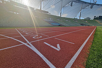 Low-angle view of the red synthetic running track at Munich Olympic Stadium in sunlight. The finish line with lane markings is clearly visible in the foreground – a key element in athletics races.