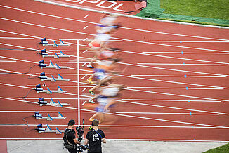 Start of a sprint final at the 2022 European Athletics Championships in Munich’s Olympic Stadium. Athletes explode from the starting blocks on the red synthetic track, captured by camera crews.