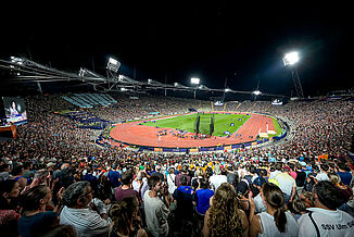 Munich Olympic Stadium during the 2022 European Athletics Championships at night. The red synthetic running track is illuminated, with thousands of spectators watching international track and field competitions under floodlights