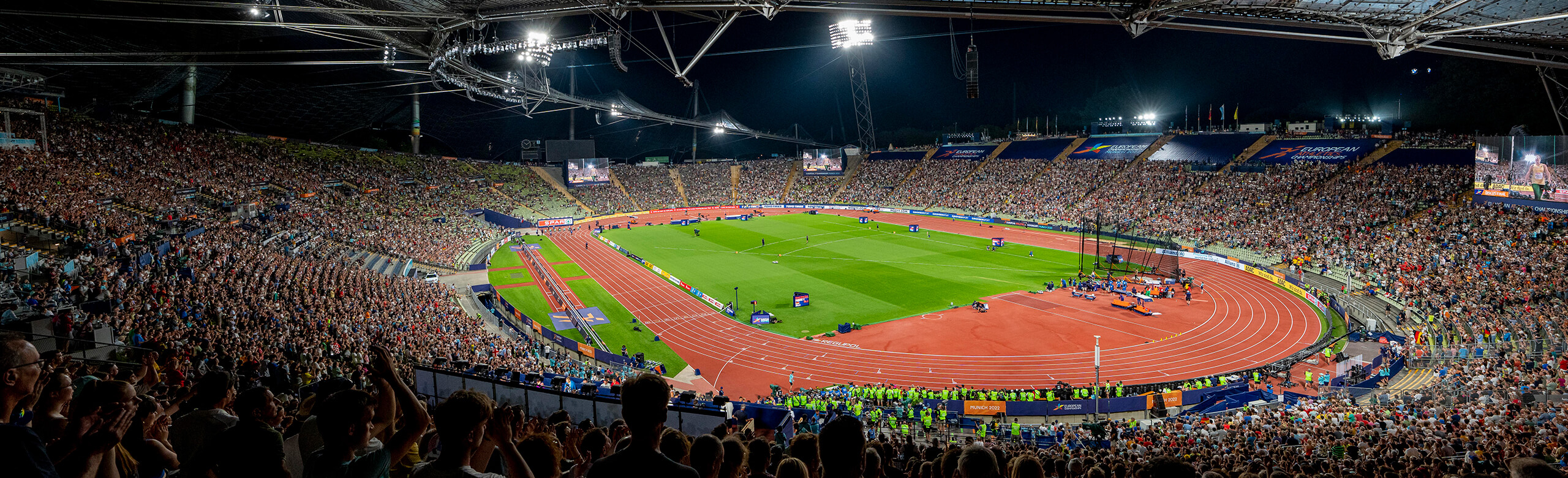 Wide-angle view of the athletics running track at Munich Olympic Stadium at night during the 2022 European Athletics Championships. The red synthetic surface is surrounded by thousands of spectators. Fully illuminated, the stadium hosts elite European track and field competition.